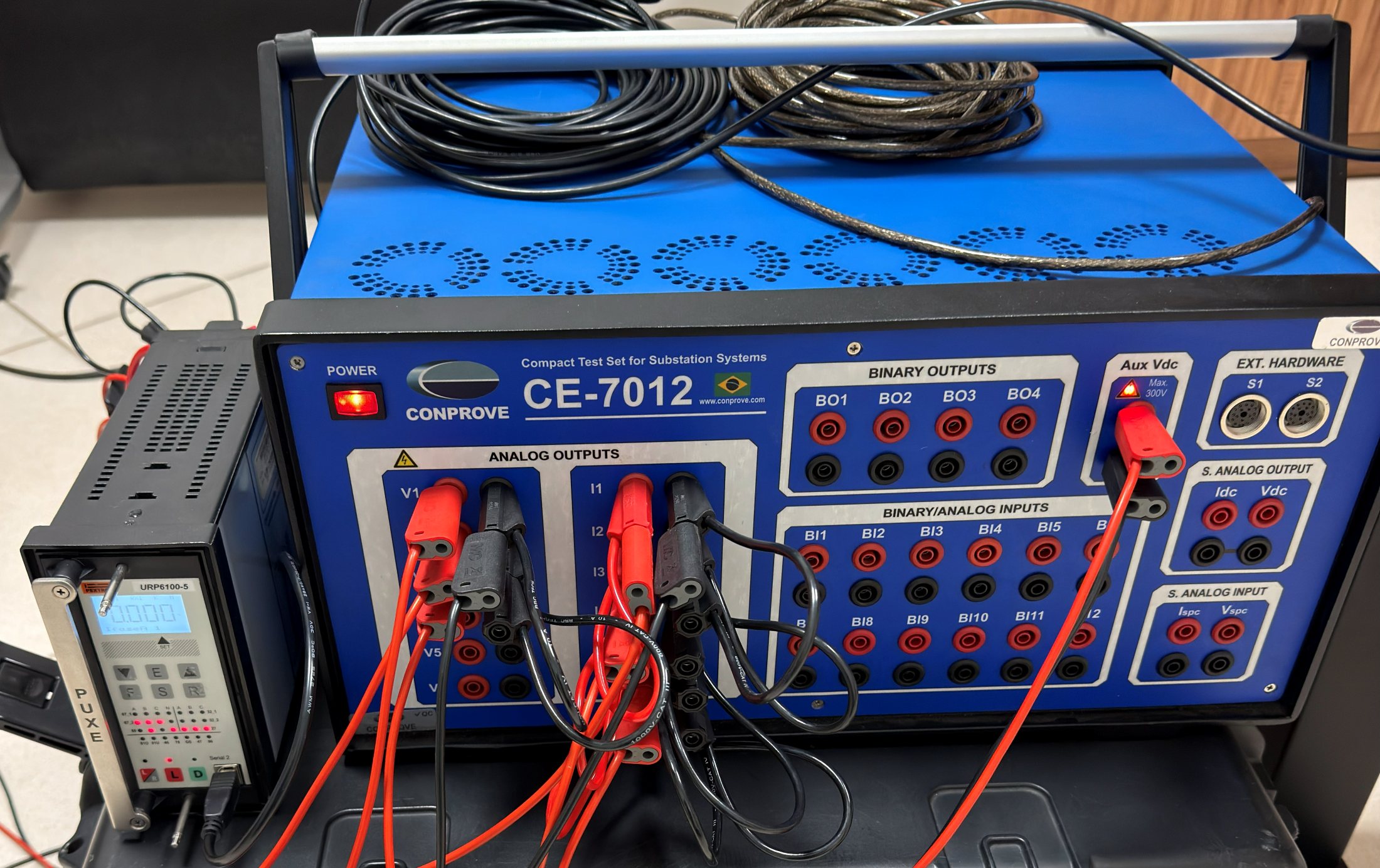 Bancada de ensaio: mala de teste Conprove CE-7012 com o relé Pextron URP6100 ao lado, ligado às saídas analógicas de tensão V1 a V3 e de corrente I1 a I3 por cabos vermelhos e pretos